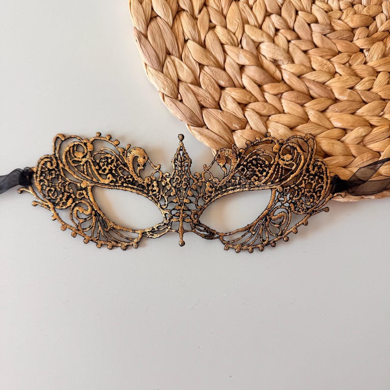 May include: Ornate black and gold masquerade mask with intricate filigree detailing. The mask features a central crown design and is attached to black ribbons. The mask is set against a neutral background with a woven texture.