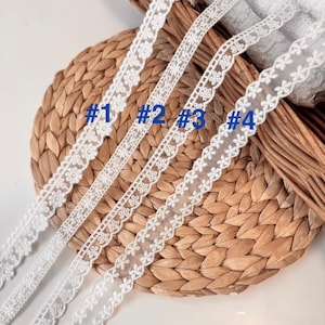 May include: Four white lace ribbons with different floral and geometric patterns are shown against a woven background. The ribbons are labelled with blue numbers: #1, #2, #3, and #4. The delicate lace is suitable for craft projects. The image shows the detail of the lace.
