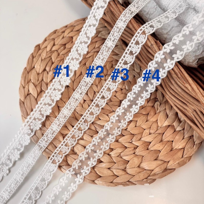 May include: Four white lace ribbons with different floral and geometric patterns are shown against a woven background. The ribbons are labelled with blue numbers: #1, #2, #3, and #4. The delicate lace is suitable for craft projects. The image shows the detail of the lace.