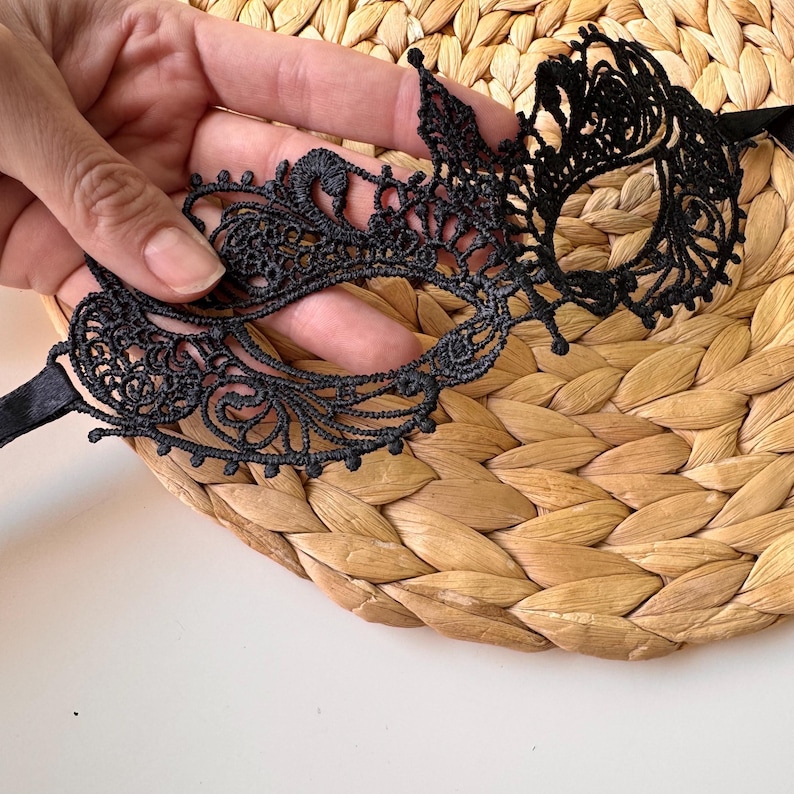 May include: A black lace eye mask with an intricate floral design. The mask is held by a hand and rests on a woven, beige surface. The mask has a satin ribbon tie.