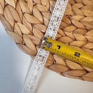 May include: Close-up of white lace ribbon with a floral pattern. A yellow measuring tape shows the ribbon is approximately 1.9 cm wide. The ribbon is placed on a woven, light brown surface.