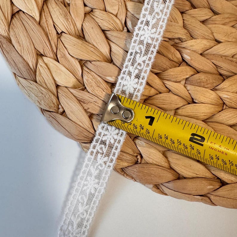 May include: Close-up of white lace ribbon with a floral pattern. A yellow measuring tape shows the ribbon is approximately 1.9 cm wide. The ribbon is placed on a woven, light brown surface.