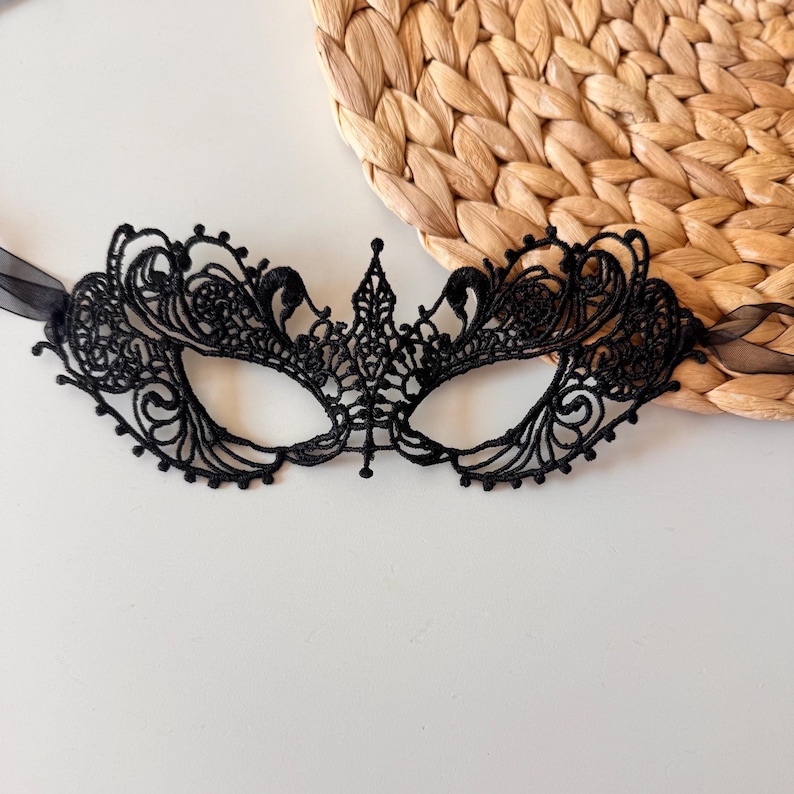 May include: Black lace masquerade mask with intricate floral and swirl patterns. The mask has a central crown-like design and is attached to black ribbons. The mask is set against a neutral background with a woven texture.