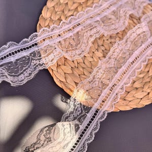 May include: Close-up of delicate white lace trim with scalloped edges and intricate floral patterns. The lace is arranged on a woven, light brown surface, creating a contrast in textures. The trim features a series of small, evenly spaced holes.