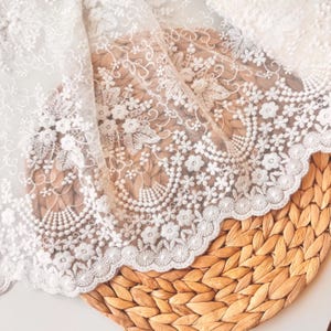 May include: Close-up of white lace fabric with floral and leaf embroidery. The lace has a scalloped edge and is draped over a woven, light brown surface. The intricate design features detailed stitching and delicate patterns.