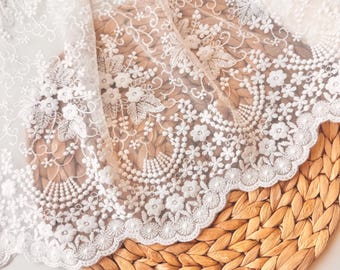 May include: Close-up of white lace fabric with floral and leaf embroidery. The lace has a scalloped edge and is draped over a woven, light brown surface. The intricate design features detailed stitching and delicate patterns.
