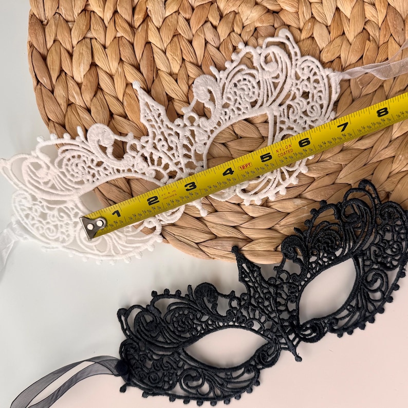 May include: Two decorative eye masks. One is white, the other is black. The white mask is measured with a tape measure at approximately 8 inches long. The masks are made of lace and are designed for a masquerade or costume event.