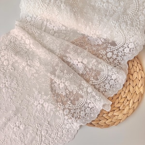 May include: White lace fabric with intricate floral and scrollwork patterns. The delicate material is folded, revealing layers of detailed embroidery. The edges are scalloped, adding a refined touch. The fabric is displayed on a woven, light brown surface.