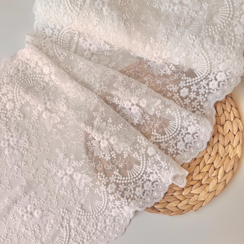 May include: White lace fabric with intricate floral and scrollwork patterns. The delicate material is folded, revealing layers of detailed embroidery. The edges are scalloped, adding a refined touch. The fabric is displayed on a woven, light brown surface.