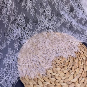 May include: White lace fabric with a delicate floral pattern. The lace has a scalloped edge and is shown on a woven basket.
