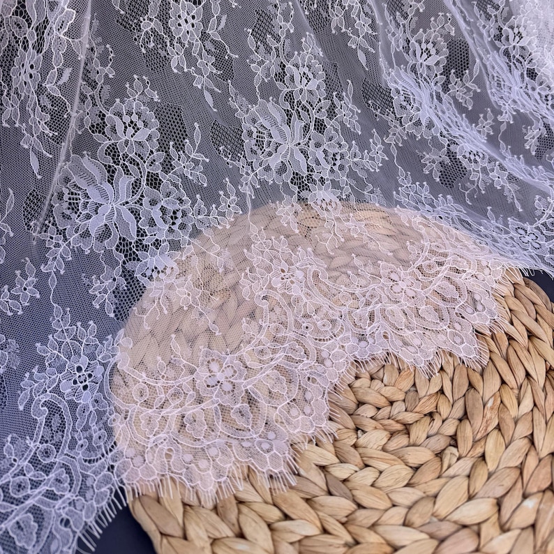May include: White lace fabric with a delicate floral pattern. The lace has a scalloped edge and is shown on a woven basket.