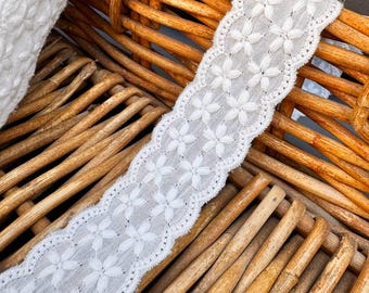 May include: White cotton lace trim with a delicate floral pattern. The lace is shown on a woven wicker basket.