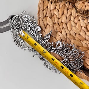 May include: A silver and black filigree masquerade mask with a ribbon tie. The mask is approximately 6 inches long and features an intricate, openwork design. The mask is displayed on a woven background.