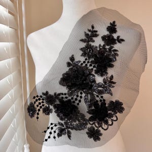 Może przedstawiać: Czarna aplikacja z koronki kwiatowej z cekinami i koralikami. Aplikacja przedstawia szczegółowy wzór kwiatów i liści, idealny do ozdabiania odzieży lub akcesoriów. Koronka jest przymocowana do przezroczystego czarnego podkładu z siatki.