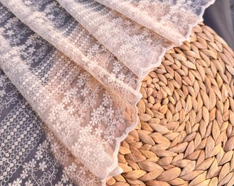 May include: Close-up of delicate, off-white lace fabric with floral and geometric patterns. The lace is draped over a woven, light brown placemat. The fabric's scalloped edge adds a soft, feminine touch to the design.