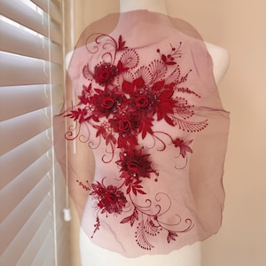 May include: A burgundy floral applique with intricate detailing. The design features multiple red roses, leaves, and delicate embellishments. The applique is on a sheer, light brown fabric, perfect for embellishing garments or accessories.