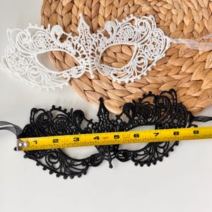 May include: Two lace masquerade masks. One is white, the other is black. The black mask is approximately 8 inches long. Both masks have intricate floral and scrollwork designs, with ribbon ties for securing them.