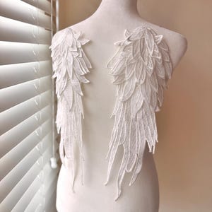 May include: A pair of white lace angel wings with a feathered design. The wings are attached to a white mannequin.