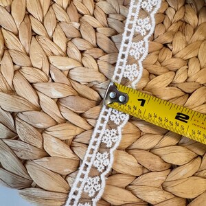 May include: White floral lace trim with a scalloped edge, measured with a yellow tape measure. The lace is approximately 2.5 cm wide. The background is a woven, natural-coloured material.