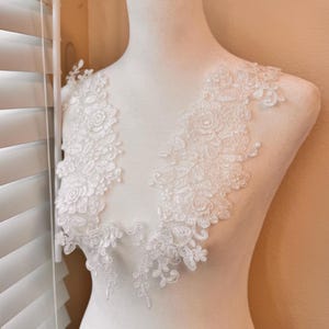 Peut inclure: Applique en dentelle blanche avec des motifs floraux et de feuilles, présenté sur un mannequin. La dentelle délicate présente des motifs détaillés de roses et d'autres fleurs, idéal pour les tenues de mariée ou de cérémonie.