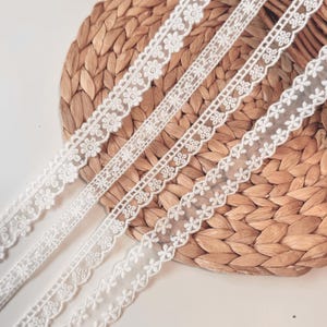 May include: Four white lace ribbons with floral and geometric designs are shown. The ribbons, varying in width, display delicate details. The lace is set against a textured, woven background, highlighting the intricate patterns and textures.