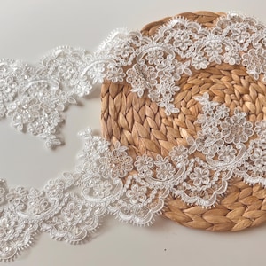 Può includere: Bordo in pizzo floreale bianco con bordo smerlato. Il pizzo presenta un design dettagliato di fiori e foglie, con piccoli abbellimenti. Il bordo è esposto su una superficie intrecciata marrone chiaro.