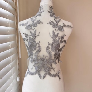 Puede incluir: Una aplicación de encaje floral gris se muestra en un maniquí. La aplicación presenta intrincados diseños florales y de hojas, con un patrón simétrico. El encaje es de color gris suave y los detalles están finamente elaborados, adecuado para ropa de novia o formal.