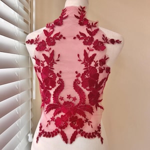 May include: A red floral lace applique with intricate embroidered details. The applique features a high neckline and a symmetrical design of flowers and leaves. The delicate lace is displayed on a mannequin, showcasing its potential for garment embellishment.