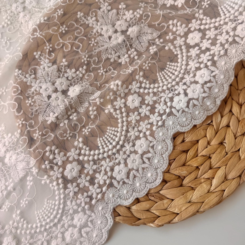 May include: Close-up of white floral lace fabric. The delicate lace features intricate embroidered flowers, leaves, and swirling patterns. The scalloped edge adds a decorative touch. The fabric is displayed on a woven, light brown surface.