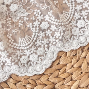May include: Close-up of white lace fabric with floral embroidery. The lace features delicate flower and leaf patterns, with a scalloped edge. The fabric is set against a woven, textured background, creating a contrast in textures.