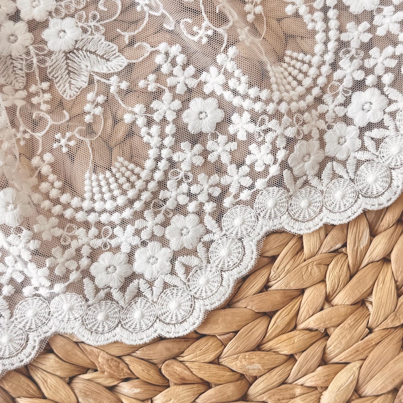 May include: Close-up of white lace fabric with floral embroidery. The lace features delicate flower and leaf patterns, with a scalloped edge. The fabric is set against a woven, textured background, creating a contrast in textures.