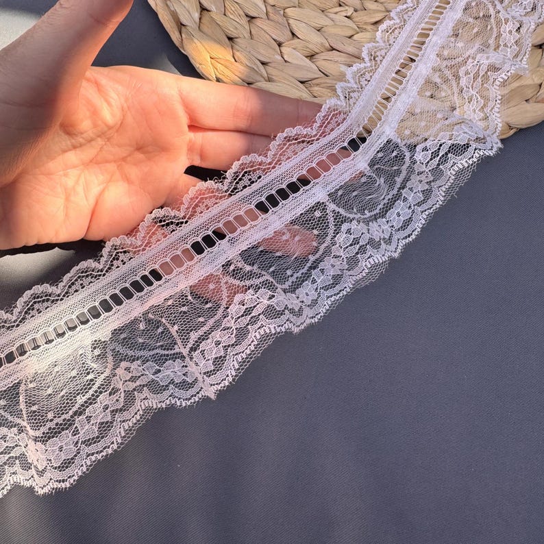 May include: Close-up of white lace trim with a delicate floral pattern and a scalloped edge. The lace features a series of rectangular cutouts along the center. The trim is on a grey surface.