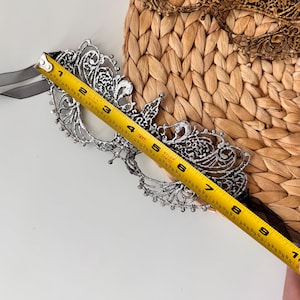 May include: A silver-colored filigree masquerade mask with a black ribbon tie. The mask is approximately 7 inches long. The mask is placed on a woven, light brown surface. A gold-colored mask is visible in the background.
