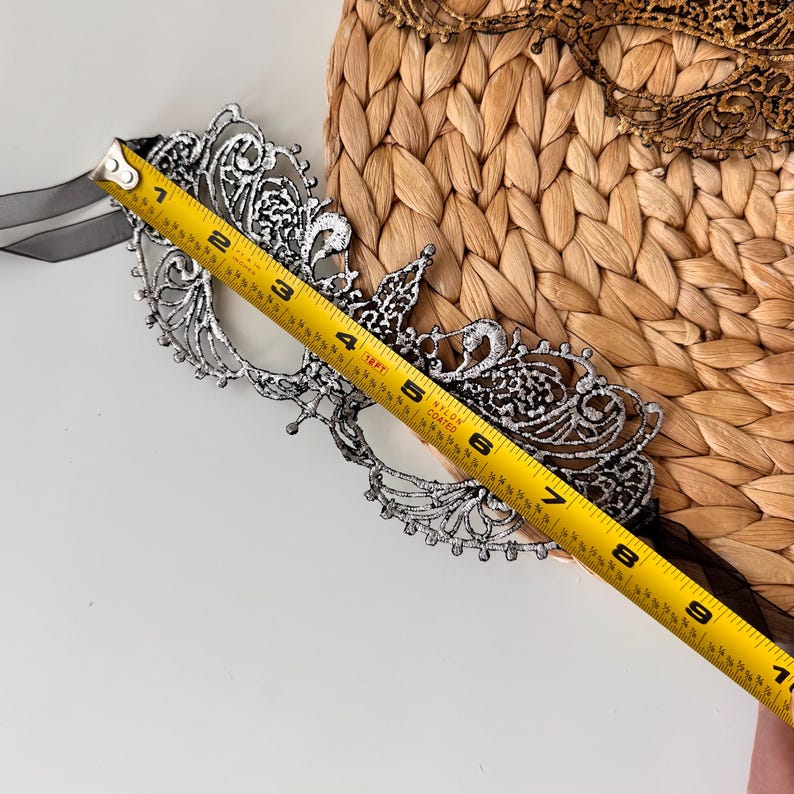May include: A silver-colored filigree masquerade mask with a black ribbon tie. The mask is approximately 7 inches long. The mask is placed on a woven, light brown surface. A gold-colored mask is visible in the background.