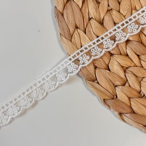 May include: A length of white lace trim with a floral design. The trim has a scalloped edge and a repeating pattern of small, delicate flowers. The lace is set against a textured, woven background.