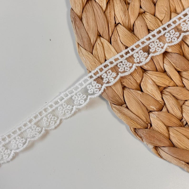 May include: A length of white lace trim with a floral design. The trim has a scalloped edge and a repeating pattern of small, delicate flowers. The lace is set against a textured, woven background.