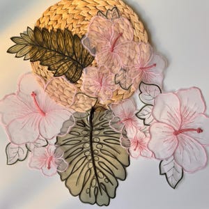 May include: A decorative floral arrangement with a woven straw base, embroidered with pink hibiscus flowers and green leaves. The arrangement includes a large, detailed leaf and smaller flower accents.