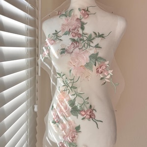 Puede incluir: Aplicación floral con flores rosas y melocotón, hojas y tallos verdes, exhibida en un maniquí. La aplicación está sobre una tela blanca transparente. Las flores están intrincadamente detalladas, creando un diseño delicado y elegante.