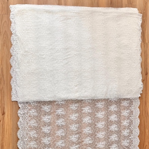 May include: White lace fabric with a floral pattern and scalloped edges. The fabric is folded, revealing a detailed texture and design. The lace is likely used for apparel or home decor projects.