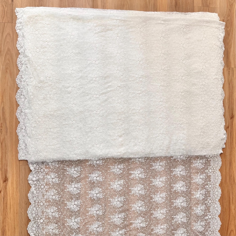 May include: White lace fabric with a floral pattern and scalloped edges. The fabric is folded, revealing a detailed texture and design. The lace is likely used for apparel or home decor projects.