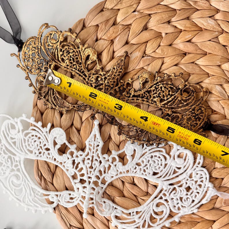 May include: A gold and black lace masquerade mask with a black ribbon tie, measuring approximately 6 inches in length, is displayed next to a white lace mask. The masks are on a woven, textured surface.