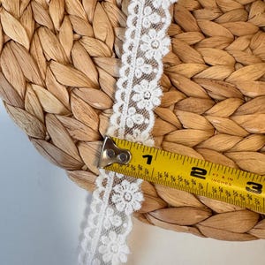 May include: White floral lace trim with a measuring tape showing a length of approximately 5 cm. The lace is placed on a woven, light brown surface. The tape measure is yellow with black markings.
