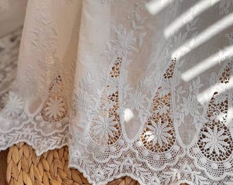 May include: White cotton fabric with a delicate floral embroidery pattern. The fabric has a scalloped edge with a lace trim.