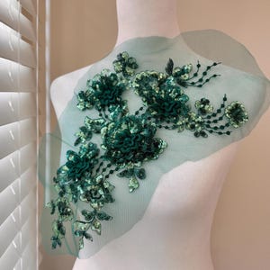 May include: Emerald green floral applique with sequins and beads. The applique features three-dimensional flower designs and delicate leaf accents, all set on a sheer, light green mesh backing. This decorative embellishment is suitable for various craft projects.