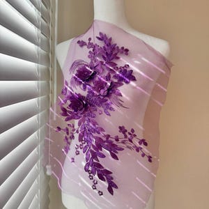 Könnte beinhalten: Lilafarbener Blumenapplikation mit Glitzerdetails auf einem durchsichtigen rosa Stoff. Die Applikation zeigt eine Ansammlung von Blumen und Blättern.