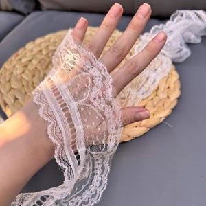 May include: Off-white lace trim featuring detailed floral and dot patterns. The lace is held up by a hand, highlighting its intricate design and texture. Suitable for sewing, crafting, or adding a vintage touch to clothing and accessories.