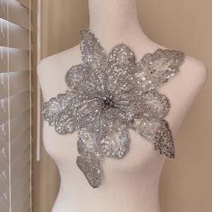 May include: A silver sequined floral applique, featuring a large, multi-petaled flower design. The applique is heavily embellished with sequins and beads, creating a shimmering effect. This decorative piece is ideal for embellishing garments or accessories.