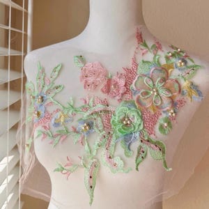 May include: A delicate floral applique with pastel pink, green, and blue flowers and leaves. The applique is adorned with small pearl-like beads and is perfect for adding a touch of elegance to clothing or crafts.