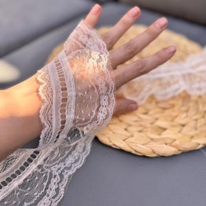May include: Elegant white lace fingerless gloves with detailed floral and dotted designs. The gloves feature scalloped edges and are partially visible against a neutral backdrop. A stylish accessory for special occasions.