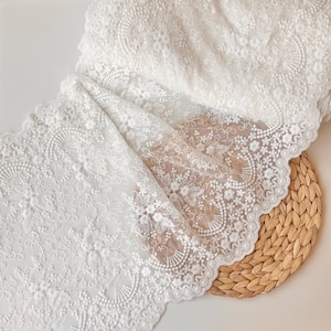 May include: White lace fabric with intricate floral and swirl patterns. The delicate material is draped over a woven, circular mat. The lace features a scalloped edge and detailed embroidery, creating a textured and elegant appearance.